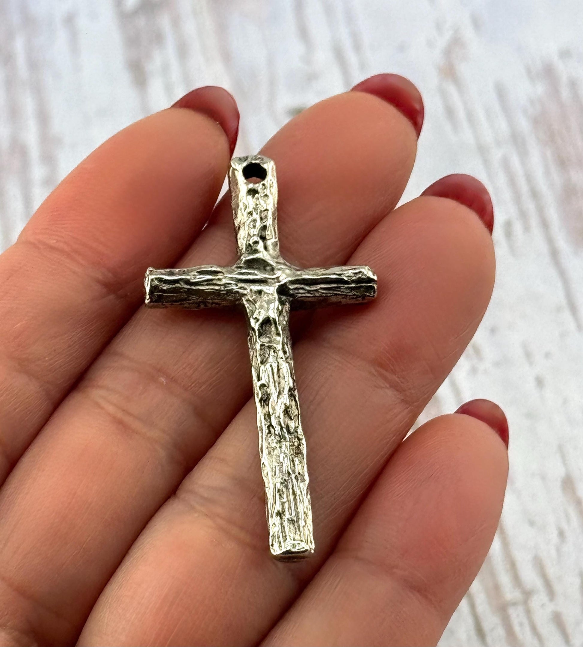 May include: A silver-colored cross pendant with a textured, bark-like appearance. The cross has a loop at the top for hanging and is held in a hand. The design is simple and rustic, suitable for religious jewelry.