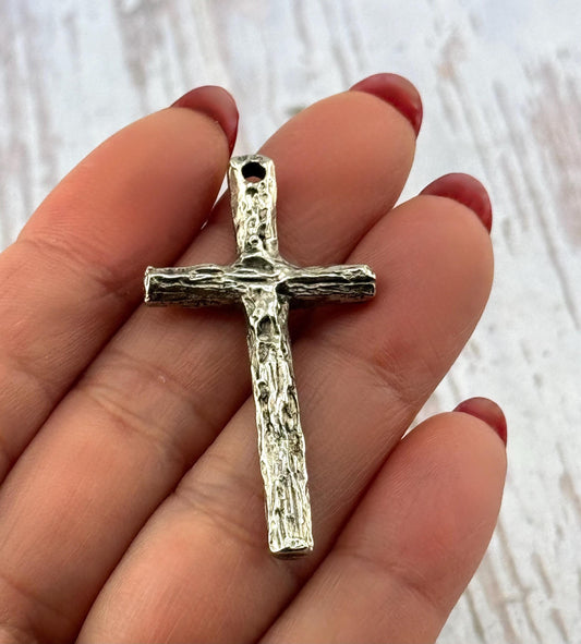 May include: A silver-colored cross pendant with a textured, bark-like appearance. The cross has a loop at the top for hanging and is held in a hand. The design is simple and rustic, suitable for religious jewelry.
